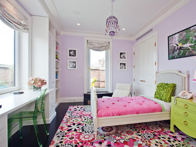 Colorful kids room in an apartment with lavender walls, a colorful rug, a chartreuse green set of drawers re purposed as a side table, a green Lucite plastic chair at a white desk with a large window, white built in bookcases and a walk in closet