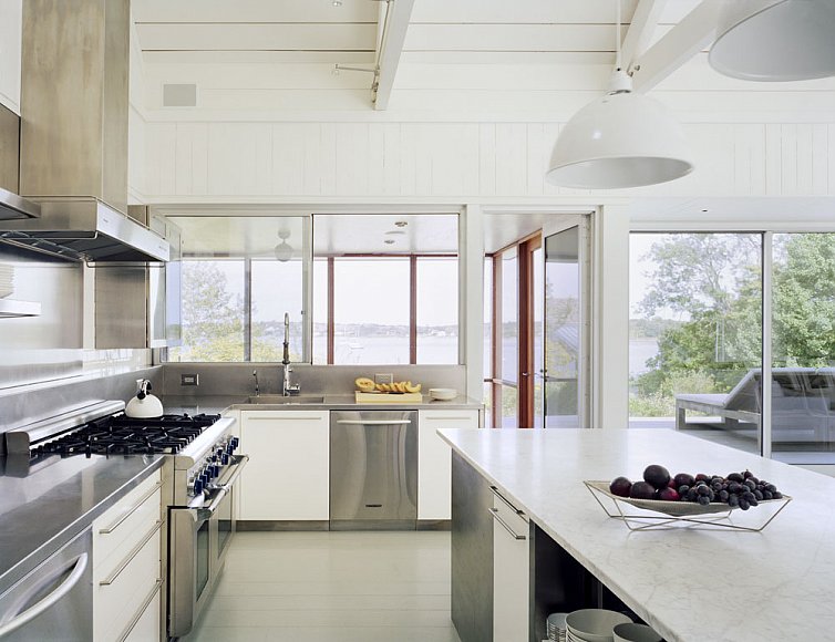 Kitchen in a Montauk on Long Island lake house with large windows, a sliding glass door, stainless appliances, white drawers with long silver drawer pulls and white pendant lights