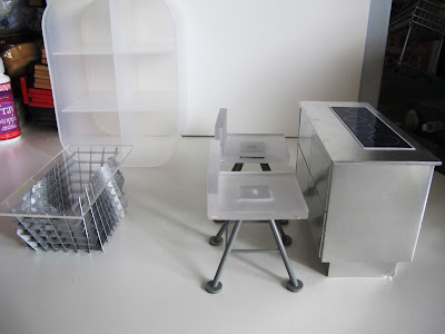 Modern dolls' house miniature set up with a metal kitchen unit with stove, white and grey bar stools, grey and perspex coffee table and a white perspex shelving unit.