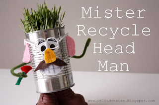 A tin can decorated as a face with grass hair, googly eyes, a felt nose, ears, and a mustache. It stands on a brown base with arms holding a red object. Text reads Mister Recycle Head Man.