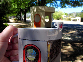 Dolls' house miniature mug with a Canberra bus shelter print, balanced on the edge of a full-sized Canberra bus shelter mug, held in front of a Canberra bus shelter.