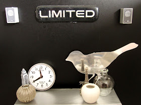 Modern dolls' house scene of a selection of grey, white and silver decoative items displayed on an industrial grey bench against a black wall. On the wall are to stereo speakers and a sign saying 'Limited'