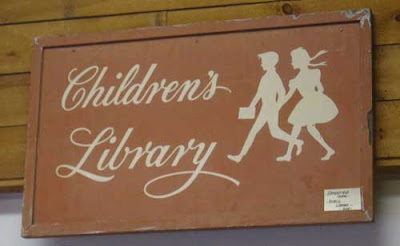 Light brown sign with hand lettered cursive Children's Library, with silhouettes of a girl and boy, probably from the 1940s