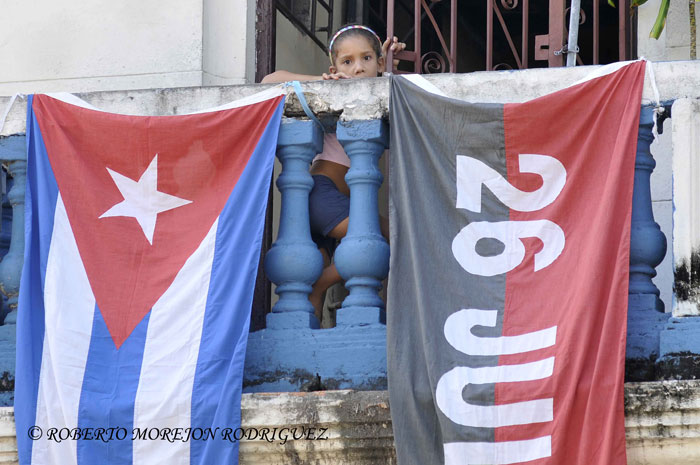 Una niña mira desde un balcón engalanado con una bandera cubana y otra del Movimiento 26 de Julio, en La Habana, Cuba, el 24 de julio de 2013. Una niña mira desde un balcón engalanado con una bandera cubana y otra del Movimiento 26 de Julio, en La Habana, Cuba, el 24 de julio de 2013.