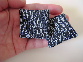 Hand holding two modern dolls' house miniature black cushions with white text on them.