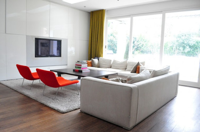living room with white sectional sofa with orange accent pillows, two red chairs, wood floor, a gray shag rug, large windows and green floor length curtains