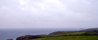 Looking across rocky fields, past cliff edges to open sea. Sky is overcast, cloudy.