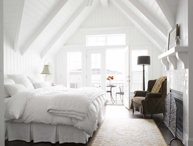 White bedroom, velvet slip covered arm chair, white bedding, white fireplace with tile chimney, French doors and an A-frame ceiling 