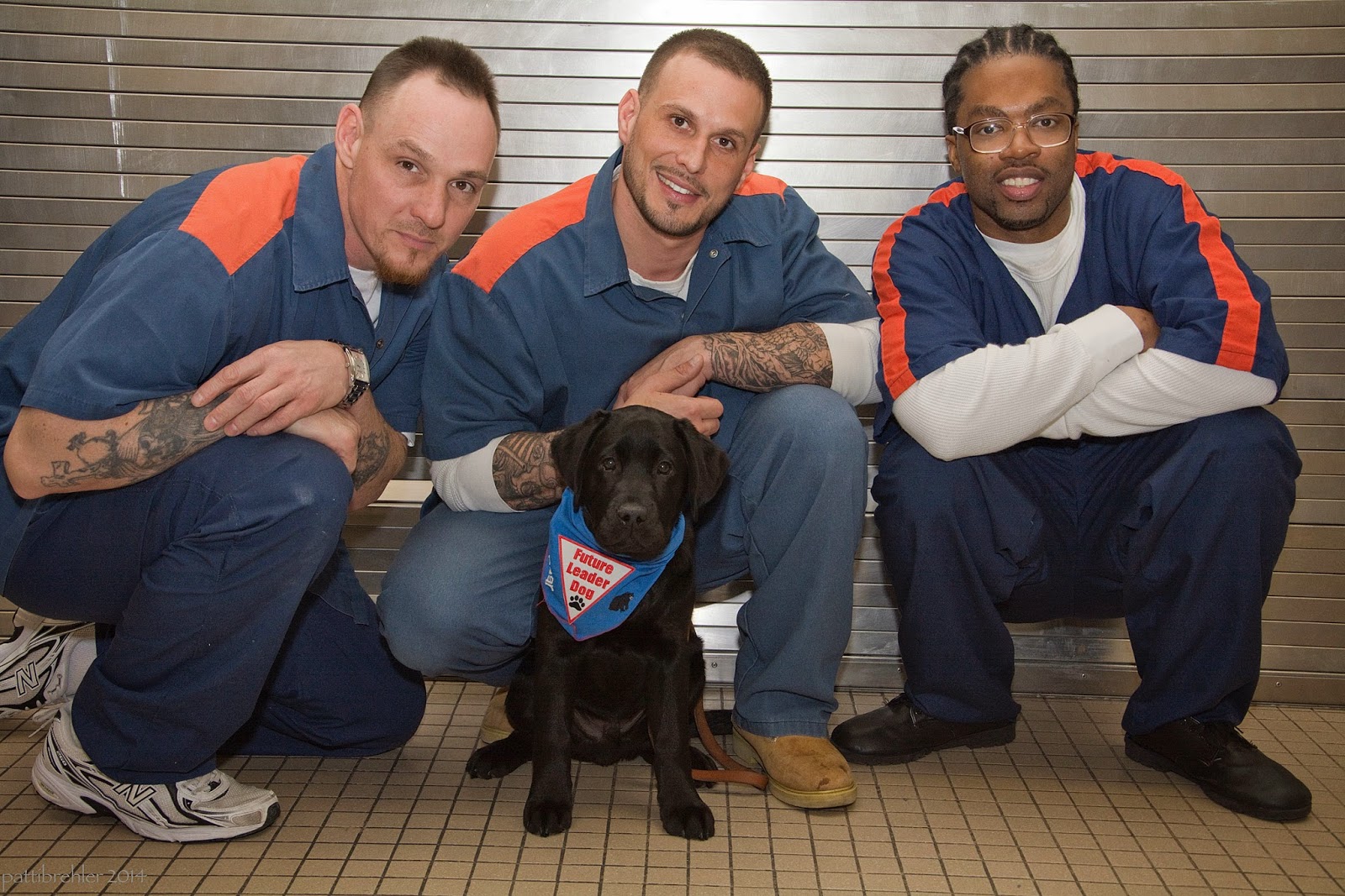 Three men are squatting in front of a steel wall on a tile floor. A young black lab puppy is sitting in front of the middle man, looking at the camera. The puppy is wearing a blue Future Leader dog bandana. The men have their arms resting on their knees and are smiling at the camera. The man on the right is an african american and is wearing glasses.