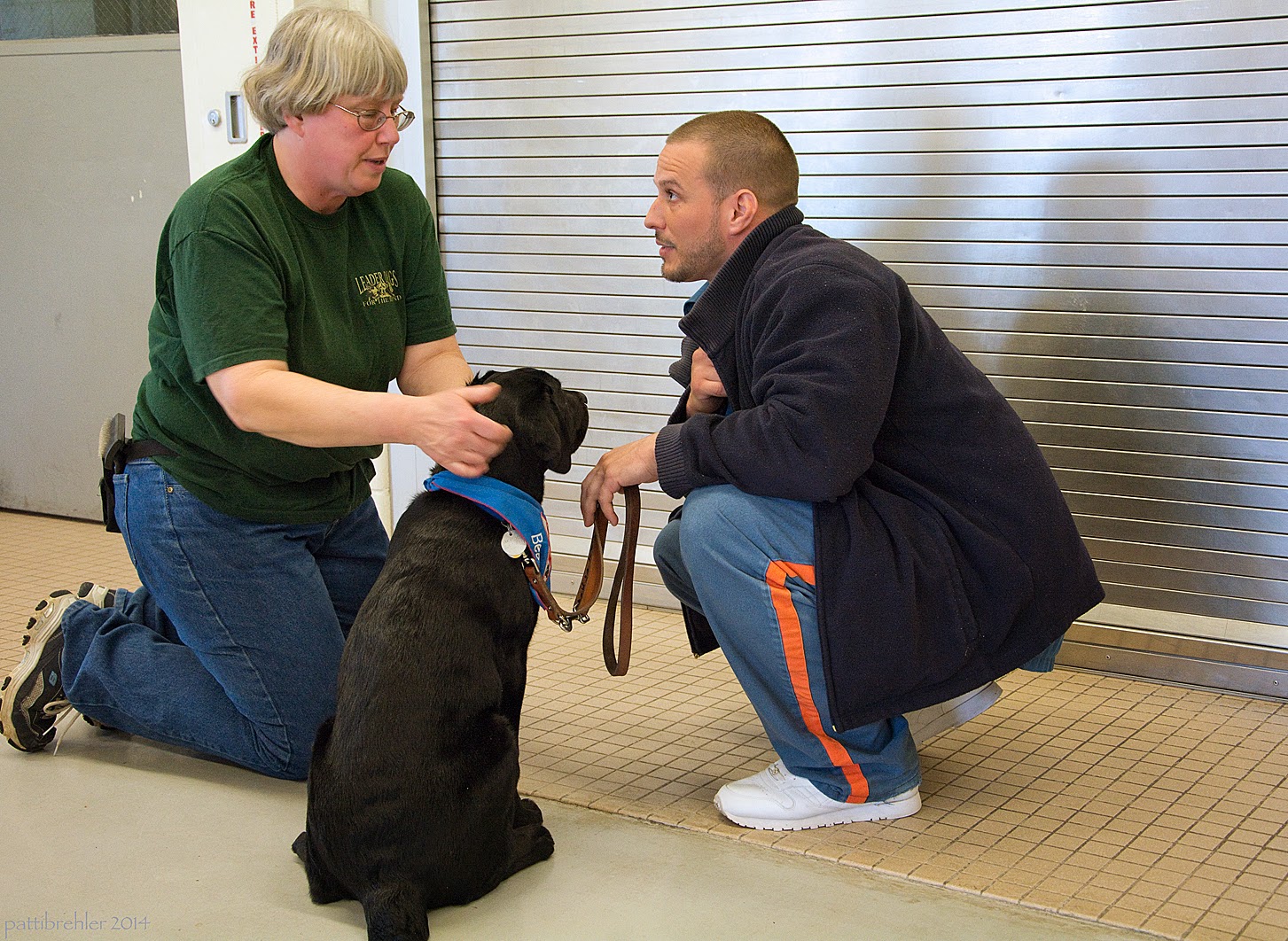 A woman dressed in a short sleeved green t-shirt and blue jeans wearing glasses and has short white hair is kneeling on the left. Her hands are checking the ears of a young black lab puppy, who is sitting on the cement floor facing away from the camera. A man is squatting to the right of the puppy. He is wearing a dark blue fleece jacket and the blue prison pants and white tennis shoes. He is holding the brown leather leash of the puppy with his left hand, which is leaning on his knee. He is looking at the woman and listening to her talk.