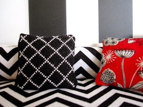 Modern miniature scene of a black and white lattice-embroidered cushion on a black and white chevron sofa against a wall with large black and white stripes. In the right corner is a red black and white mid-century modern flowered cushion.