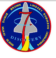 Blue back ground with outline of space shuttles and large red numeral 7 and names of crew members around the circumference.