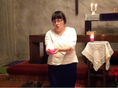 We see a small portion of a medieval chapel still in modern use. We see part of the altar, laid with a purple cloth, two candles and a Godly Play Risen Christ figure. In front of the altar rail is a woman holding a short pink pillar candle, which is lit. To her left and behind her is a tiny table covered in a white linen and lace tablecloth, on which one lit purple pillar candle remains.