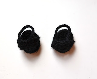 A pair of black crocheted sandals. The sole is oval shaped to match the bottom of the foot. The foot slips into a strap over the instep and another strap secures the sandal around the ankle.