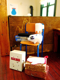 Selection of modern dolls house miniatures including a school chair with blue legs, an old first aid cupboard, two school cases, games of snakes and ladders and checkers, a blue and white piggy bank and a red checked flask.