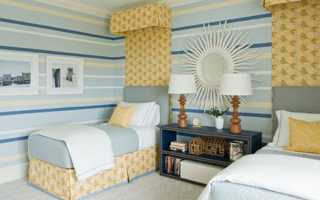 Striped walls in shades of blue and yellow coordinate with simple blue upholstered headbaords with a yellow floral crown and drape that match  the pleated bedskirts in a twin bedroom