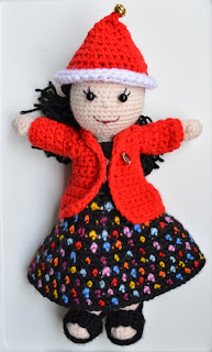 Kwokkie Doll is wearing a red santa's hat with white trim and a jingle bell on the top.  She is wearing a black skirt which has multi-coloured polka-dots matched with a black sleeveless top covered with a red jacket. On her feet, Kwokkie Doll is wearing black crocheted sandals.