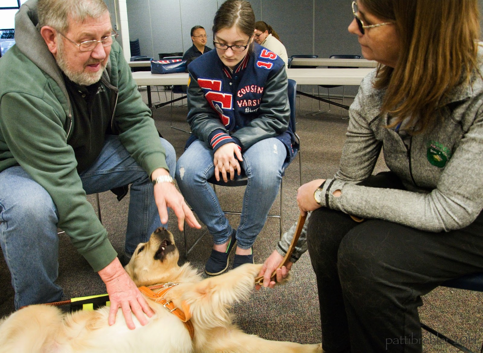 The man in the green sweatshirt is sitting on a chair on the left side, reaching to pet the golden retriever. The golden is lying on his side on the carpet, rolling over. Next to the man is a teenage girl with glasses and a varsity jacket and blue jeans. She is sitting in a chair looking down at the dog at her feet. On the right side is the woman with sunglasses wearing a grey shirt and black slacks. She is holding the dog's leash in her hands.