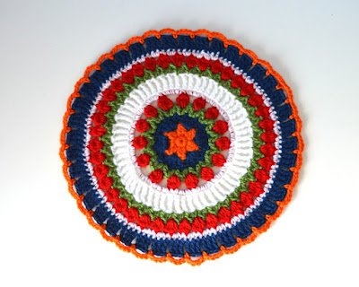 Mandala has an orange star in the centre surrounded by a blue circle. Two concentric rings of green 'leaves' and red 'tulip flowers' are separated by a wider ring of tall white stitches. After the second ring of tulips is a thin stripe of white double crochets, ending with a wider blue border with orange chain loops to finish. 