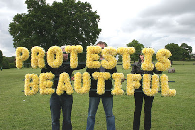photo of three people holding letters made of yellow flowers that spell protest is beautiful