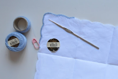 A scalloped handkerchief designed for crocheting - it is already finished off with holes around the edges, ready to insert stitches.  I have worked blue double crochet stitches around the corner and the work is secured with a pink locking stitch marker.  The 0.75 mm steel hook is laying on top of the handkerchief next to the round label from the cotton reel.  Two cotton reels are alongside the handkerchief and viewed from above. The size 80 thread is Anchor brand, colour 129.