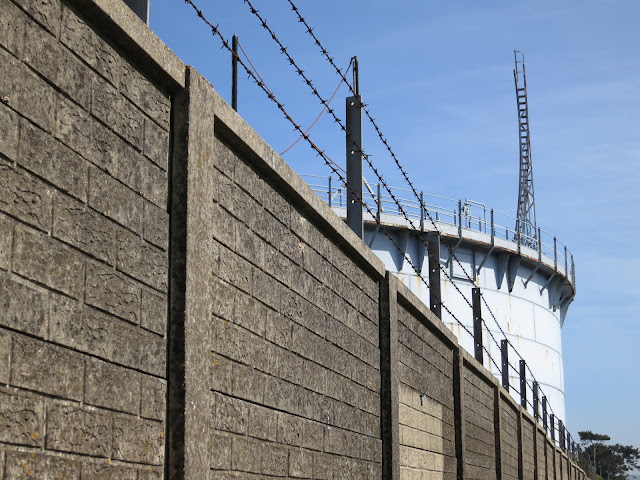 Long grey wall, topped by barbed wire, behind which is lowered gasometer.