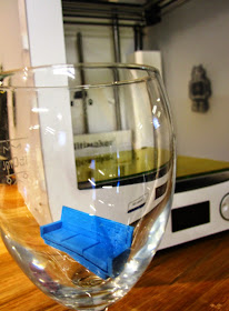 Miniature 3D-printed sofa in an empty wine glass in front of the 3D printer that printed it.