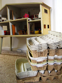 A stack of empty egg cartons in front of a vintage Lundby dolls' house.