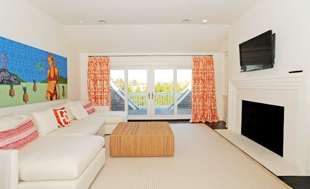 den with white walls and matching rug, dark wood floor, a fireplace, wall mounted tv, floor length orange curtains and a wooden ottoman