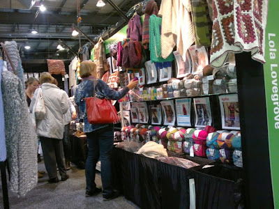 The back wall of Lola Lovegrove has two rows of patterns on display and underneath each pattern is the yarn for that pattern. Across the top of the stand are handcrafted garment samples.  In the top right corner one can see a crocheted granny square blanket.