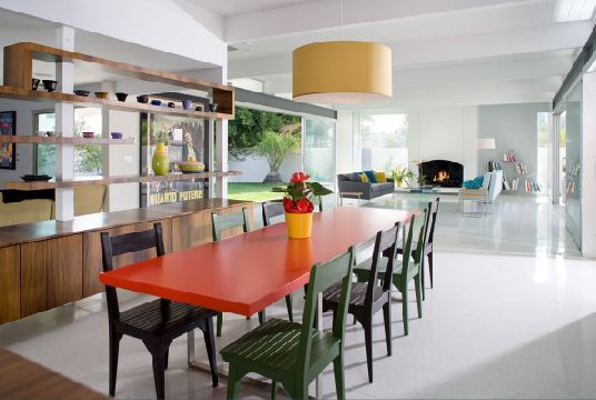 Dining room with an oval yellow pendant light, red table, green and black chairs, a wood floor and a white area rug