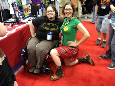 Author cosplaying Oracle, using blue wheelchair, wearing black Batman logo shirt with brown cargo pants & headset, posed with Jennie Breeden wearing red plaid kilt, green shirt, & funky boots