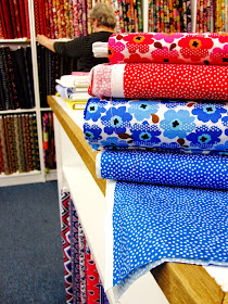 Inside a quilting fabric shop, showing a pile of fabric with Marimekko-like mini prints in the foreground and someone choosing fabric from the shelf in the background.