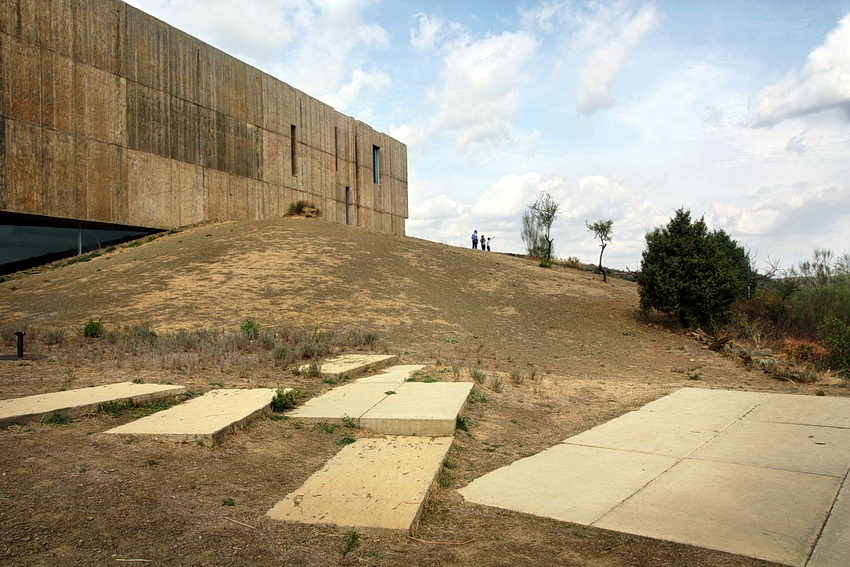 Foto com o edifício à esquerda, pessoas ao longe e palcas de cimento em primeiro plano