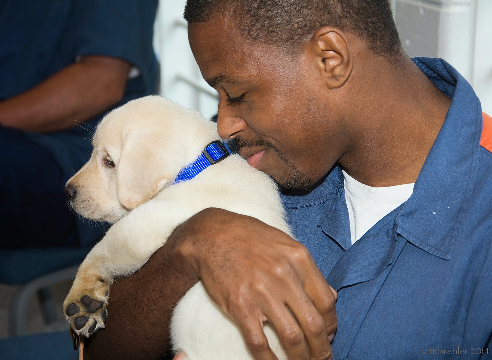An african american man dressed in a blue prison shirt is pressing his face against a small yellow lab puppy that he is holding with his right arm. The man is facing the right, and he has a contented smile. The puppy is also facing right with its left paw hanging over the man's forearm.
