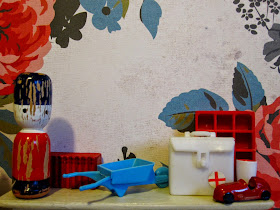 Selection of vintage miniature accessories displayed on a dolls' house mantlepiece. Colours are predominantly red, cream and mid=blue.