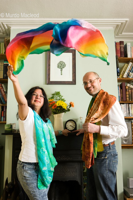 Neelam throws a LitB scarf into the air Neelam & Andrew (the LitB directors) are standing either side of a fireplace. Neelam is throwing a colourful scarf into the air