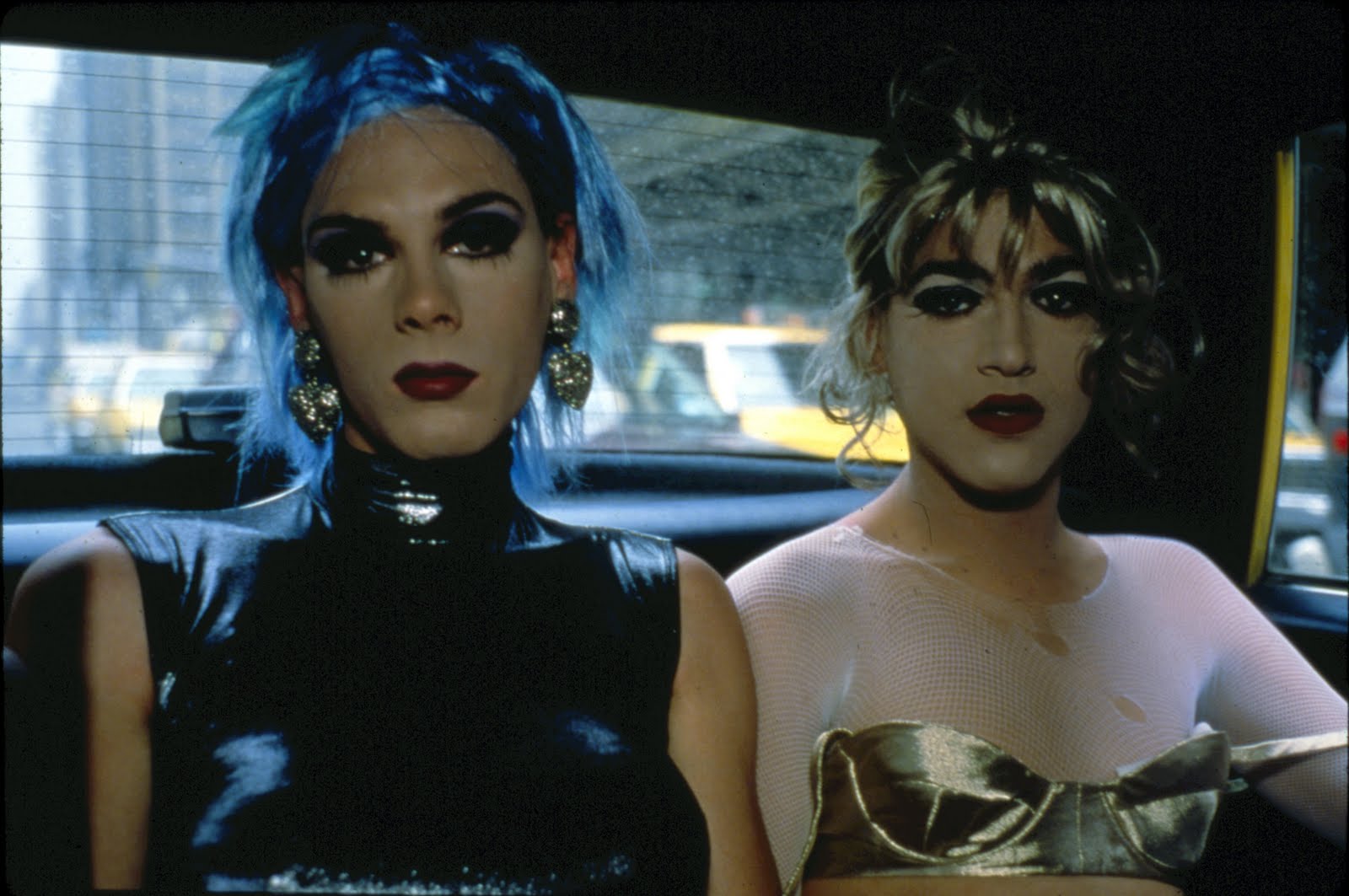 A couple of drag queens are looking directly to the camera, they seem to be in a cab