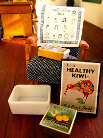 Collection of miniature items including a 1950s-style chair, a tea towel, a stack of old National Geographic magazines, a white plastic tub, a 'Be a health Kiwi' poster and an old biscuit tin.