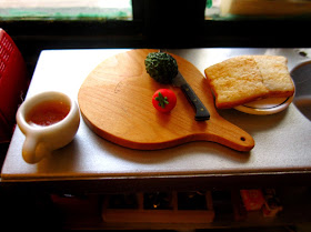 Modern dolls' house miniature stainless steel bench with a chopping board with a knife, an avocado and a tomato on it. Next to it is a plate of toast and a mug of tea.