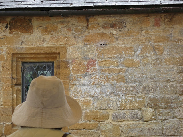 Woman with brown hat sits in front of brown stone wall with window.