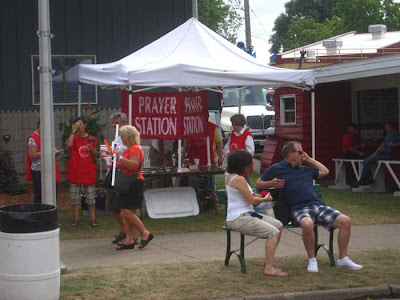 Large red banner with white letters reading Prayer Station, volunteers in red tshirts around it