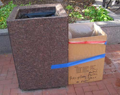 Stone trash container with cardboard box taped to it with blue tape, labeled Recycling