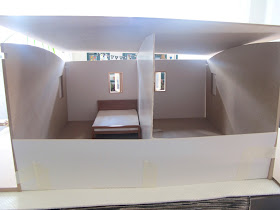 Front view of a taped-together modern dolls' house miniature kit, with a cardboard half-wall and two small rooms, one with a bed in it.