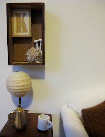 Modern dolls' house miniature scene of a side table with lamp, coffee pot and mug. On the wall above it is a box frame with the letter K, a ball of wool and two knitting needles.