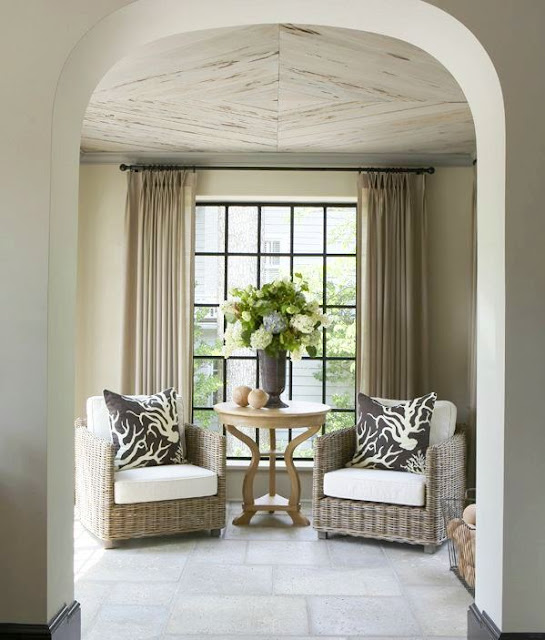 Wicker chairs in a sitting area in front of window with black trim with a large bouquet of hydrangeas in a vase on a round wooden table