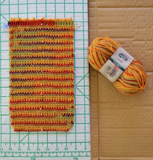 A rectangular shaped bag, flat on the blocking board with its yarn skein alongside. The yarn is variegated which produces stripes on the bag.