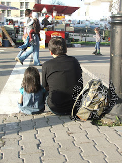 járdaszegélyen ül egy apa a lányával, miközben előttük emberek járkálnak, gyermekek görkoriznak az úton. a távolban pattogtatott kukorica árus bódéja látható.
