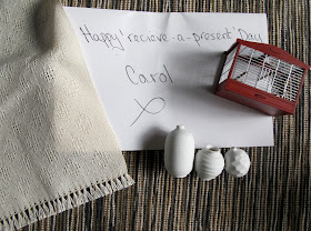 A card on which is written 'Happy receive-a-present Day', surrounded by five modern dolls' house miniature items: a hand-woven bedspread, three white pottery vases and a birdcage.