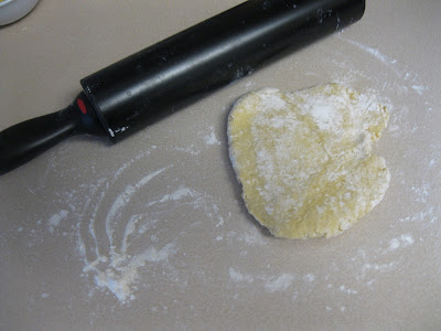 A flattened piece of dough dusted with flour rests on a beige countertop next to a black rolling pin. White flour is scattered around the area, indicating a baking preparation in progress.